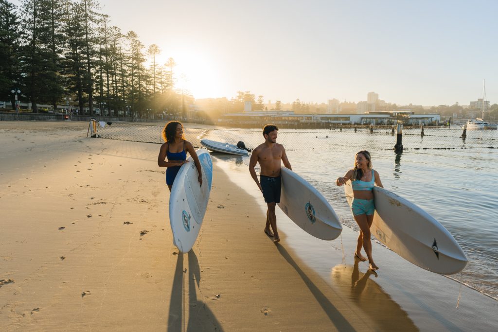 3 people carry stand up paddleboards along Manly Cove
